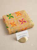 A folded beige embroidered fabric piece with colorful floral motifs in orange, green, and pink, displayed on a textured beige surface next to a small white card reading MOHAN TEXTILES.