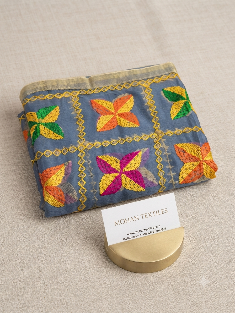 A folded piece of blue fabric with colorful embroidered butterfly motifs and gold geometric borders, displayed on a beige textured surface with a small MOHAN TEXTILES card in front.