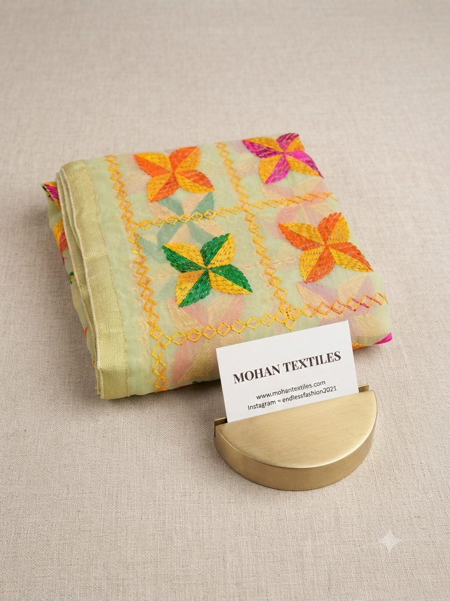 A folded light green textile with colorful embroidered floral motifs in yellow, orange, pink, and green, displayed on a beige textured surface with a small gold stand holding a business card for Mohan Textiles.