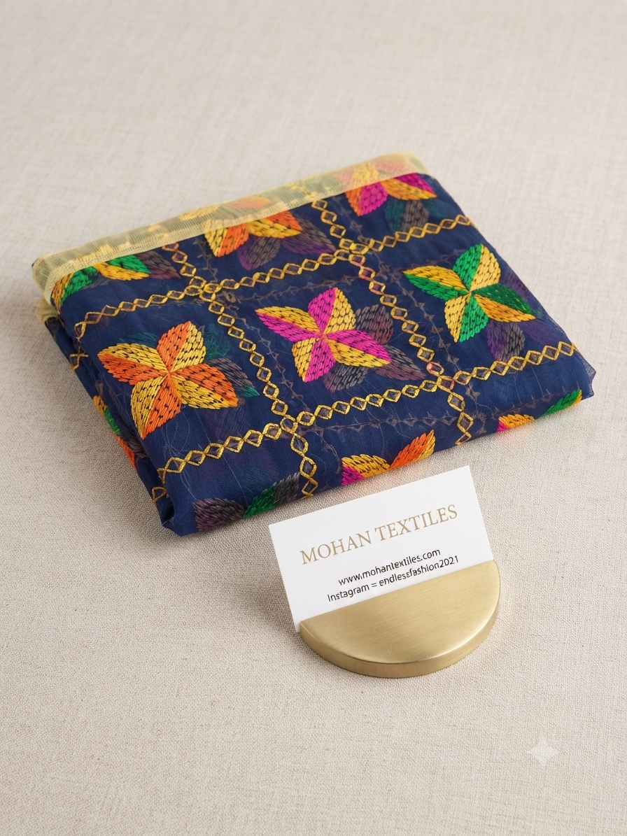 A folded navy blue textile with colorful butterfly motifs and gold chain patterns, displayed on a neutral beige surface next to a white business card for Mohan Textiles.