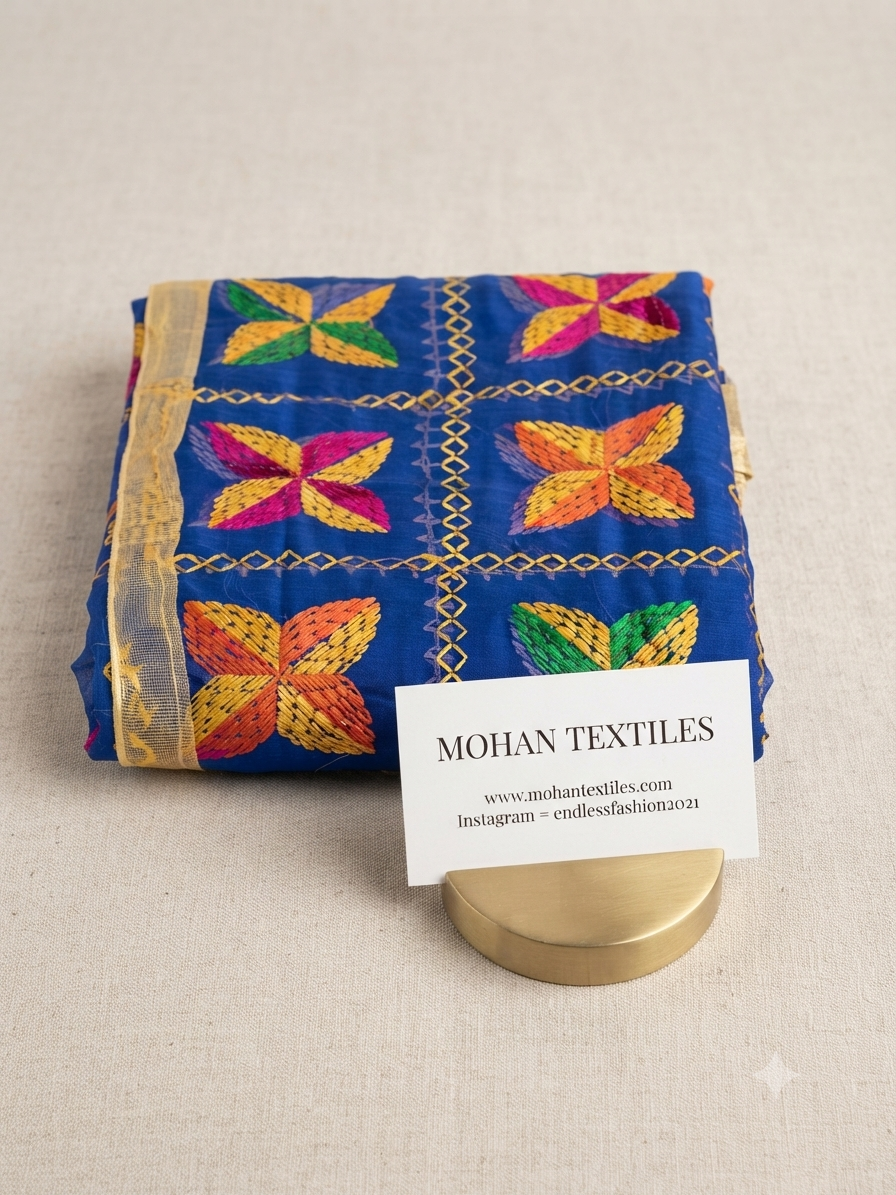A folded blue fabric piece with colorful embroidered floral motifs and gold trim, displayed on a beige textured surface with a small white card reading MOHAN TEXTILES and a gold stand.