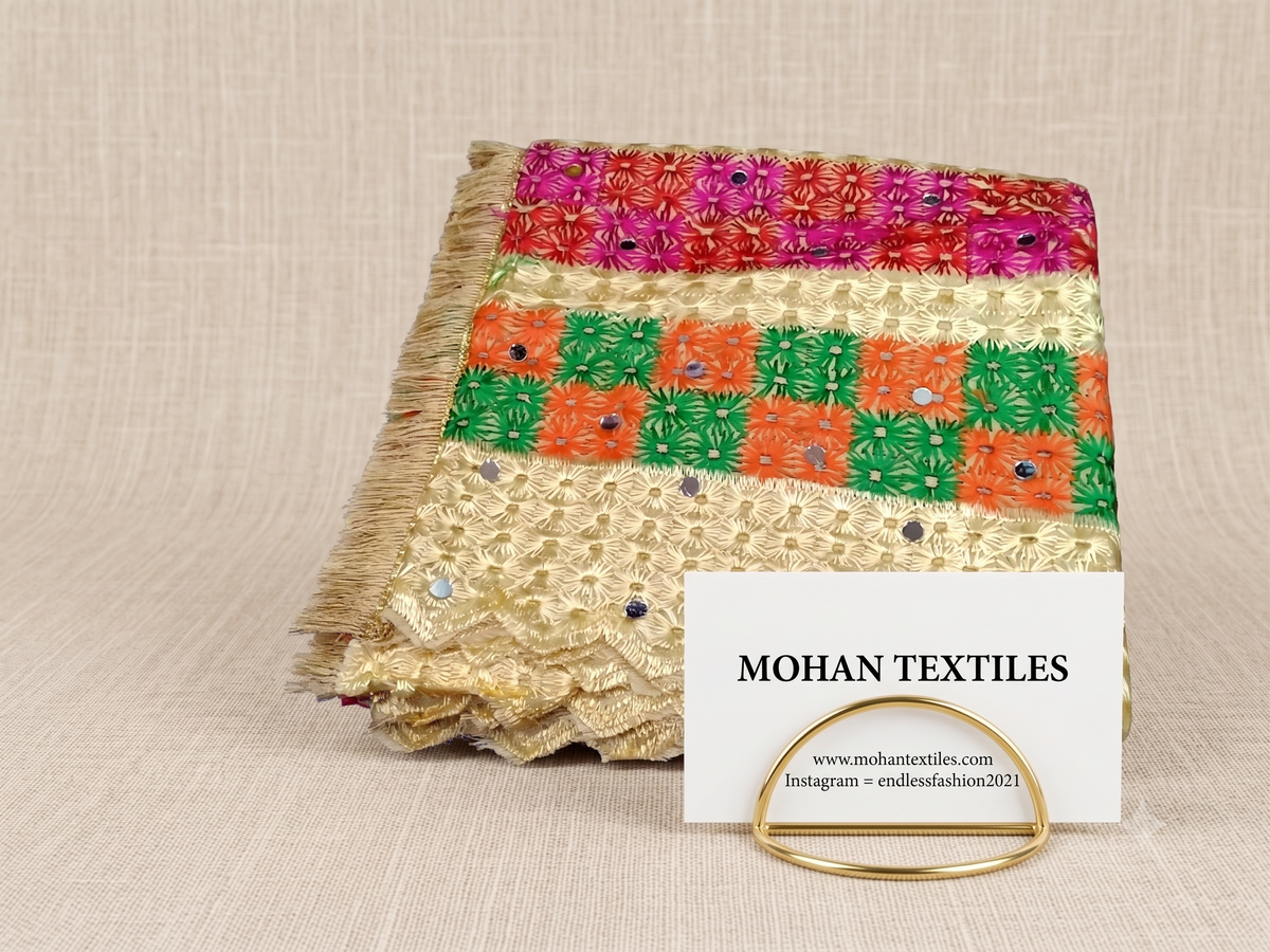 A folded piece of phulkari dupatta  of Mohan Textiles with horizontal stripes of pink, green, orange, and gold floral patterns, displayed on a neutral beige textured background with a white brand card.