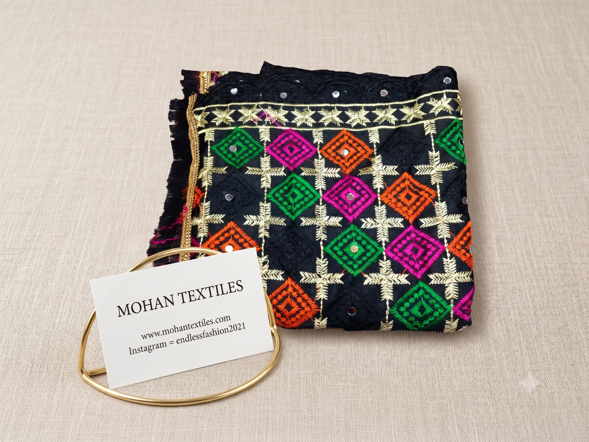 A folded black phulkari embroidered dupatta with colorful geometric patterns in green, pink, orange, and gold, displayed on a beige textured background with a gold bangle and a white brand tag reading 'MOHAN TEXTILES'.