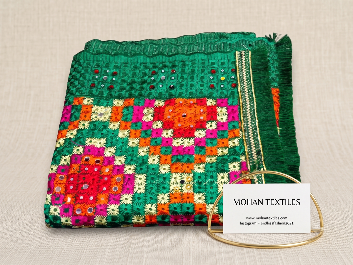 A folded green phulkari embroidered dupatta with colorful geometric patterns in pink, orange, and yellow, decorated with small mirror embellishments. A white card reading MOHAN TEXTILES is attached with a gold metal loop on a neutral beige fabric background.