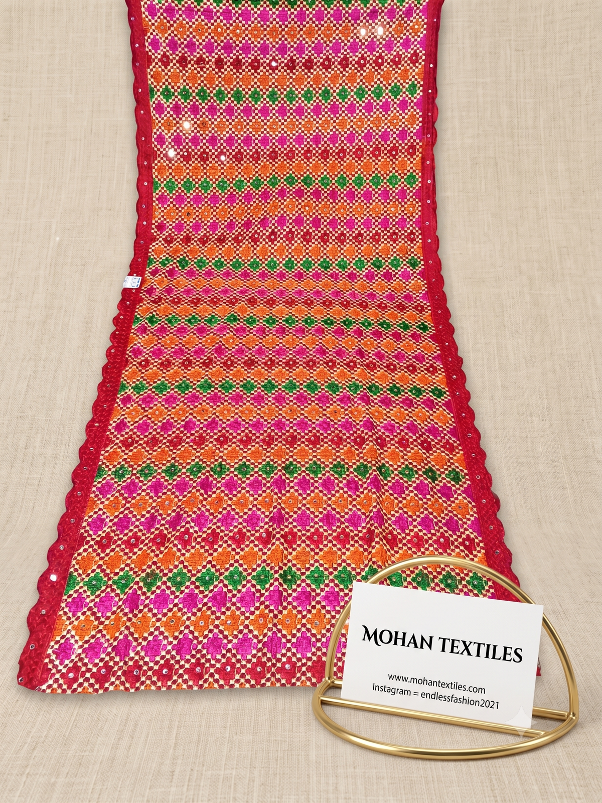 A close-up of a colorful woven Phulkari Dupatta laid flat on a beige fabric background, with a small gold stand holding a white label that reads MOHAN TEXTILES. The textile has bright pink, orange, green, and white striped woven patterning with red scalloped edges.