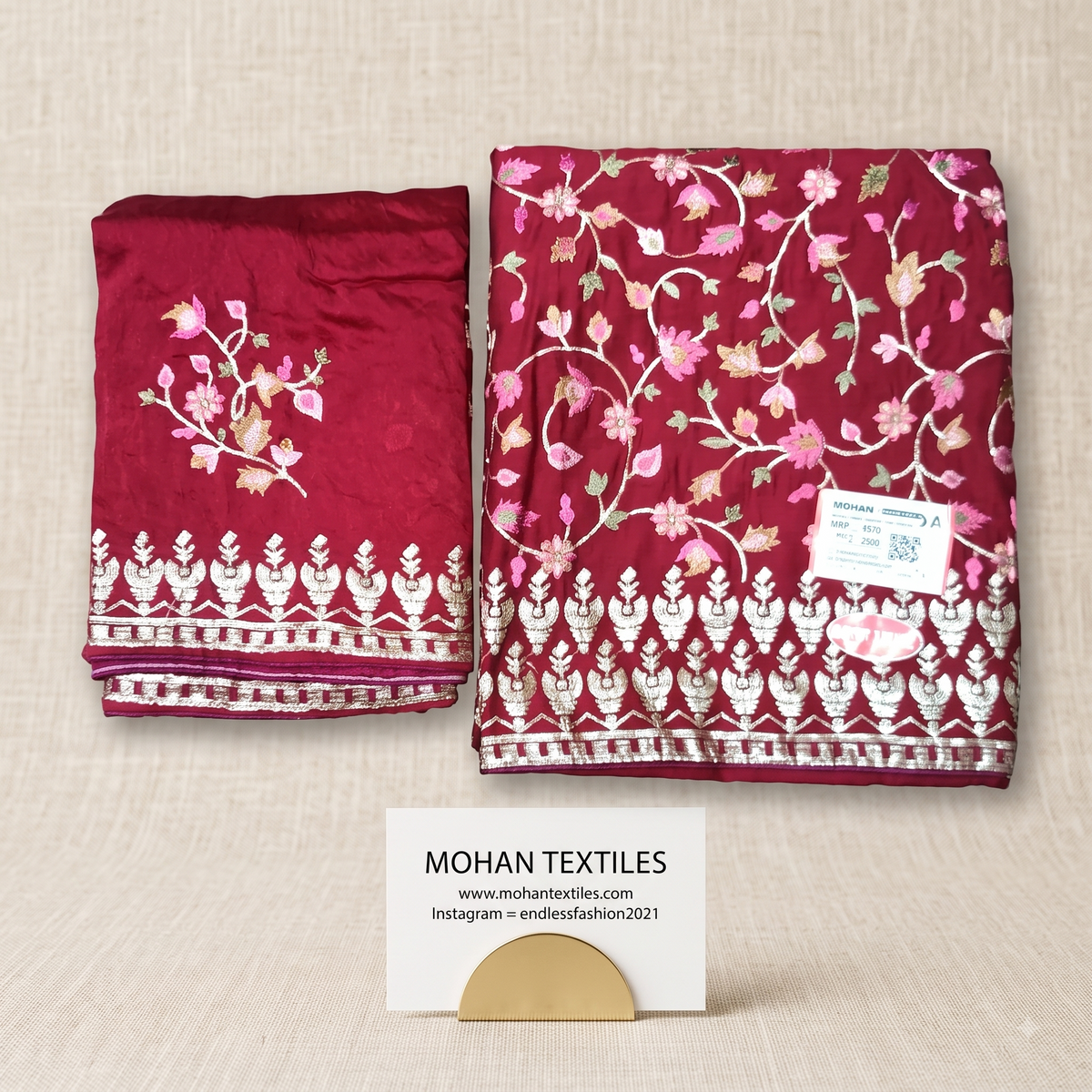 An unstitched Amritsari Suit folded deep red fabric set with pink and white floral embroidery and a white decorative border, displayed on a beige textured background. A small white card at the bottom reads MOHAN TEXTILES.