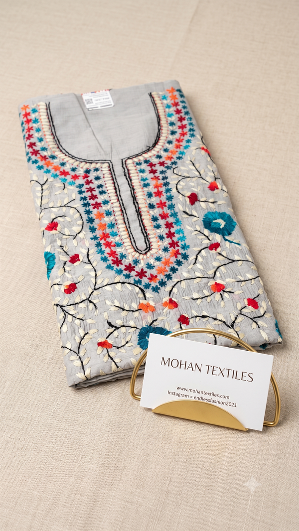 A folded light gray phulkari kurti fabric with colorful embroidered floral and vine patterns in blue, red, orange, and black, displayed on a beige textured surface with a gold ring and a card reading MOHAN TEXTILES.