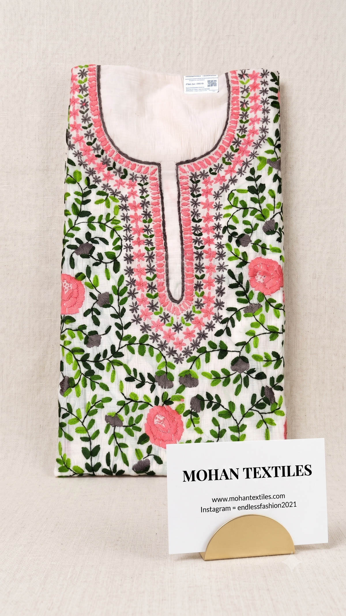 A folded white kurta with pink floral embroidery, green leafy vines, and black berry details, displayed on a beige fabric background with a brand card for Mohan Textiles.