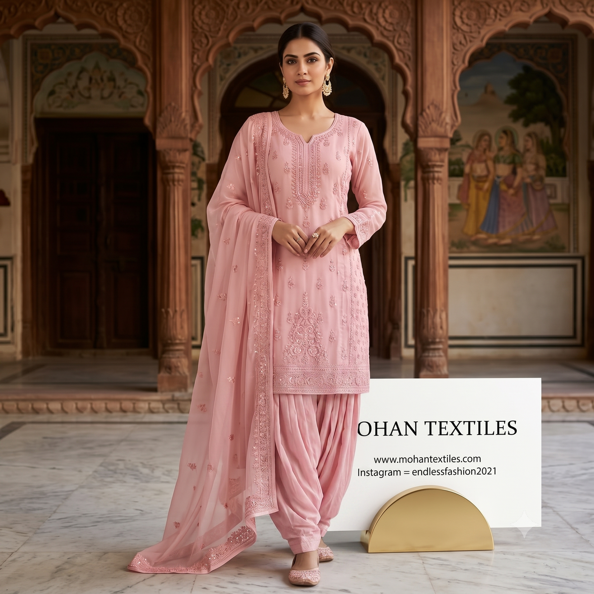 A woman models a light pink traditional Indian salwar kameez suit with matching dupatta in an ornate courtyard. The set features a pink embroidered kurta, matching pink pants, and a sheer pink dupatta.