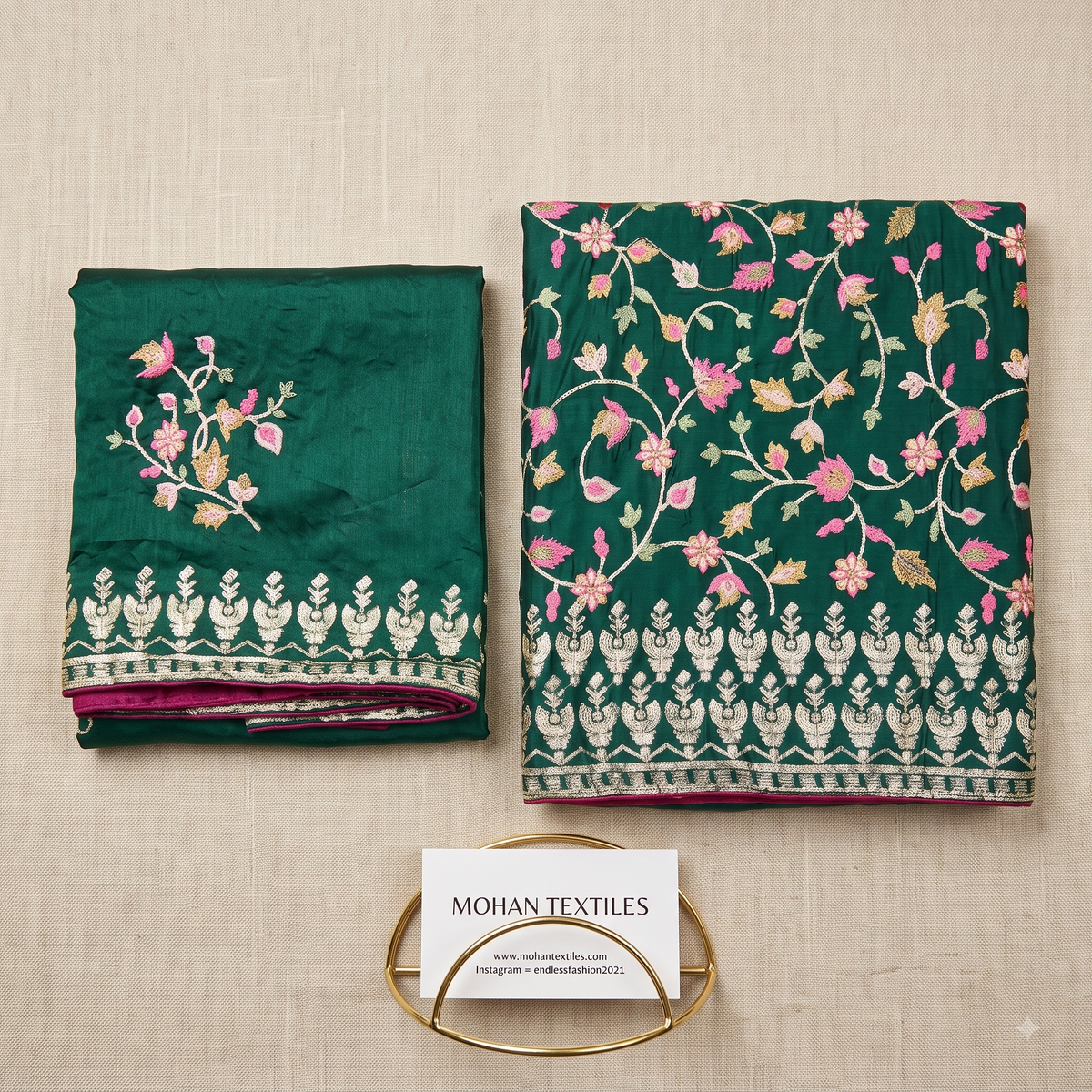 A folded and flat-laid dark green punjabi unstitched dress material set with pink and gold floral embroidery and a white border print, displayed on a light beige textured background. A small card reading MOHAN TEXTILES is placed below the fabric.