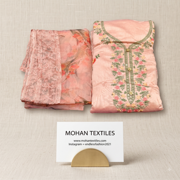 A folded peach-pink fabric set displayed on a light beige textured surface, including a floral printed fabric piece with lace trim and a matching embroidered blouse-front fabric panel with gold-tone buttons. A small card at the bottom reads MOHAN TEXTILES with website and Instagram details.