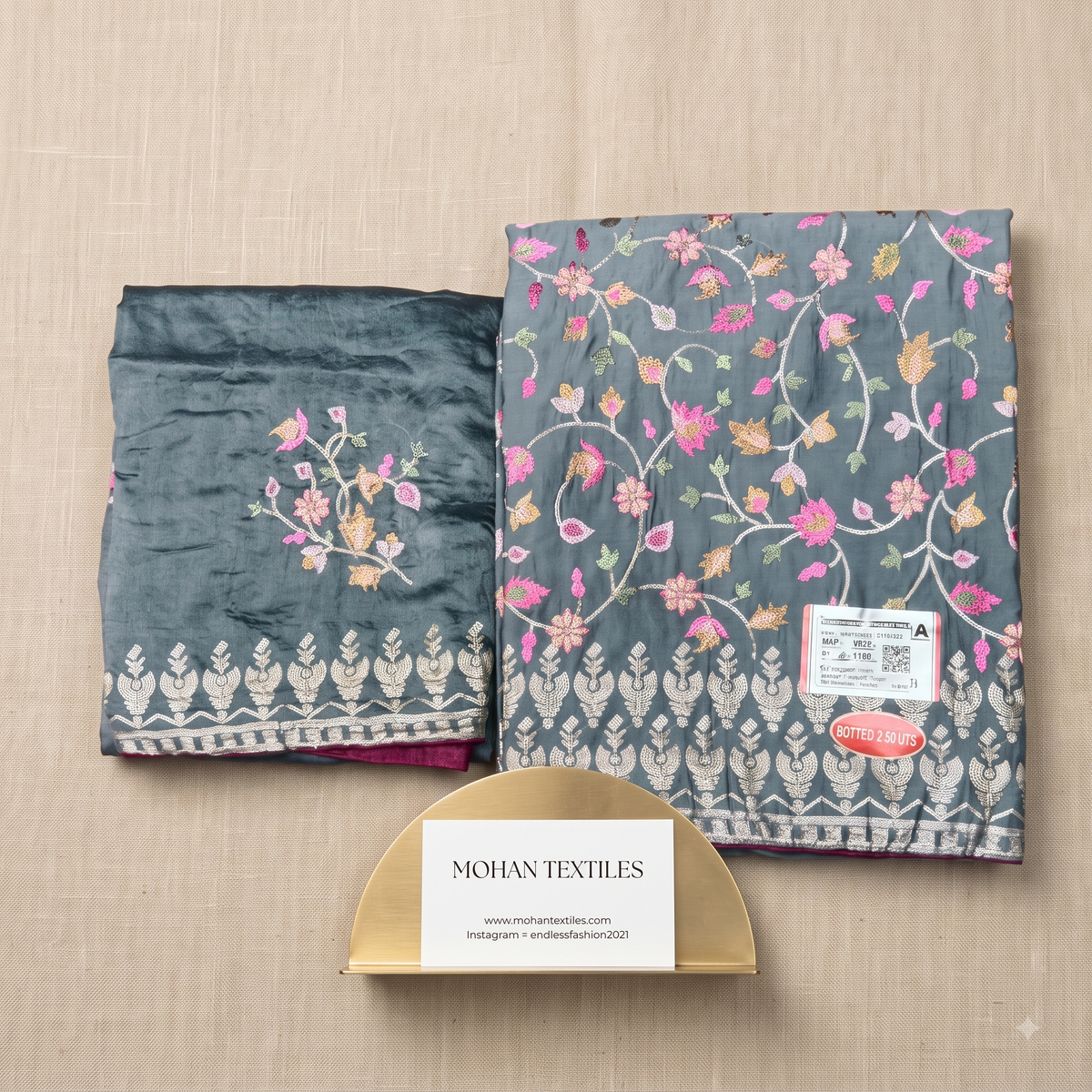 Unstitched Punjabi Dress Material laid flat on a beige textured surface, with a small gold stand in front. The fabric appears to be a dark blue-gray or slate tone with pink, peach, and cream floral/vine embroidery and a white geometric border.