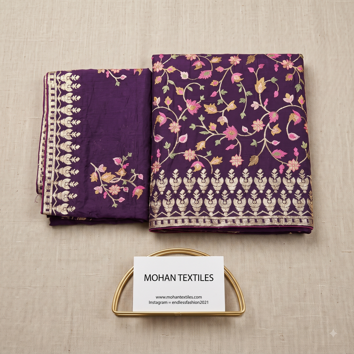 A folded piece of purple fabric with floral vine embroidery and a decorative border, displayed on a beige textured surface with a small card reading MOHAN TEXTILES.