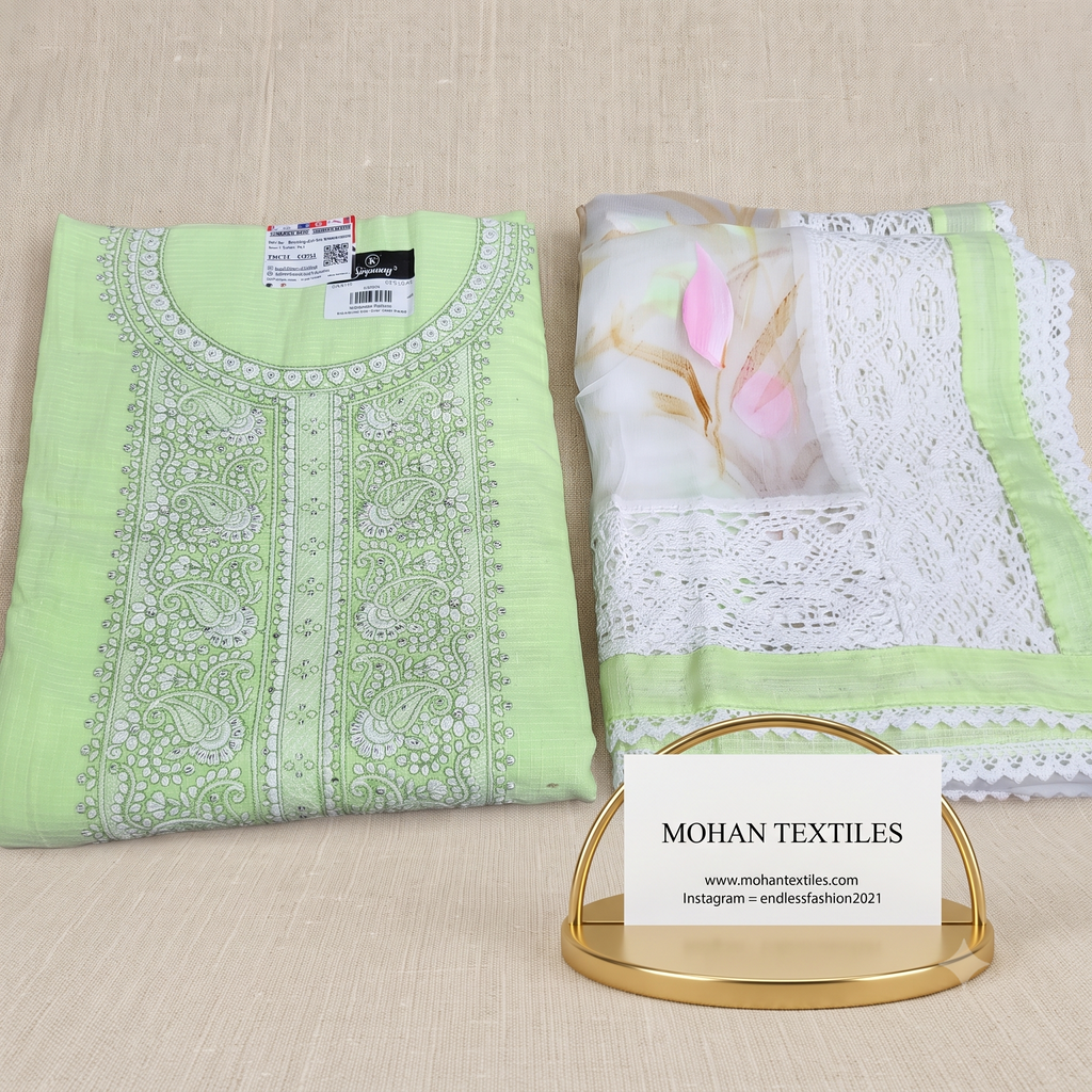 A folded light green karachi style chickenkari unstitched suit set next to a matching sheer white dupatta with floral print and lace border, arranged on a beige textured surface. A small gold stand holds a card reading MOHAN TEXTILES.
