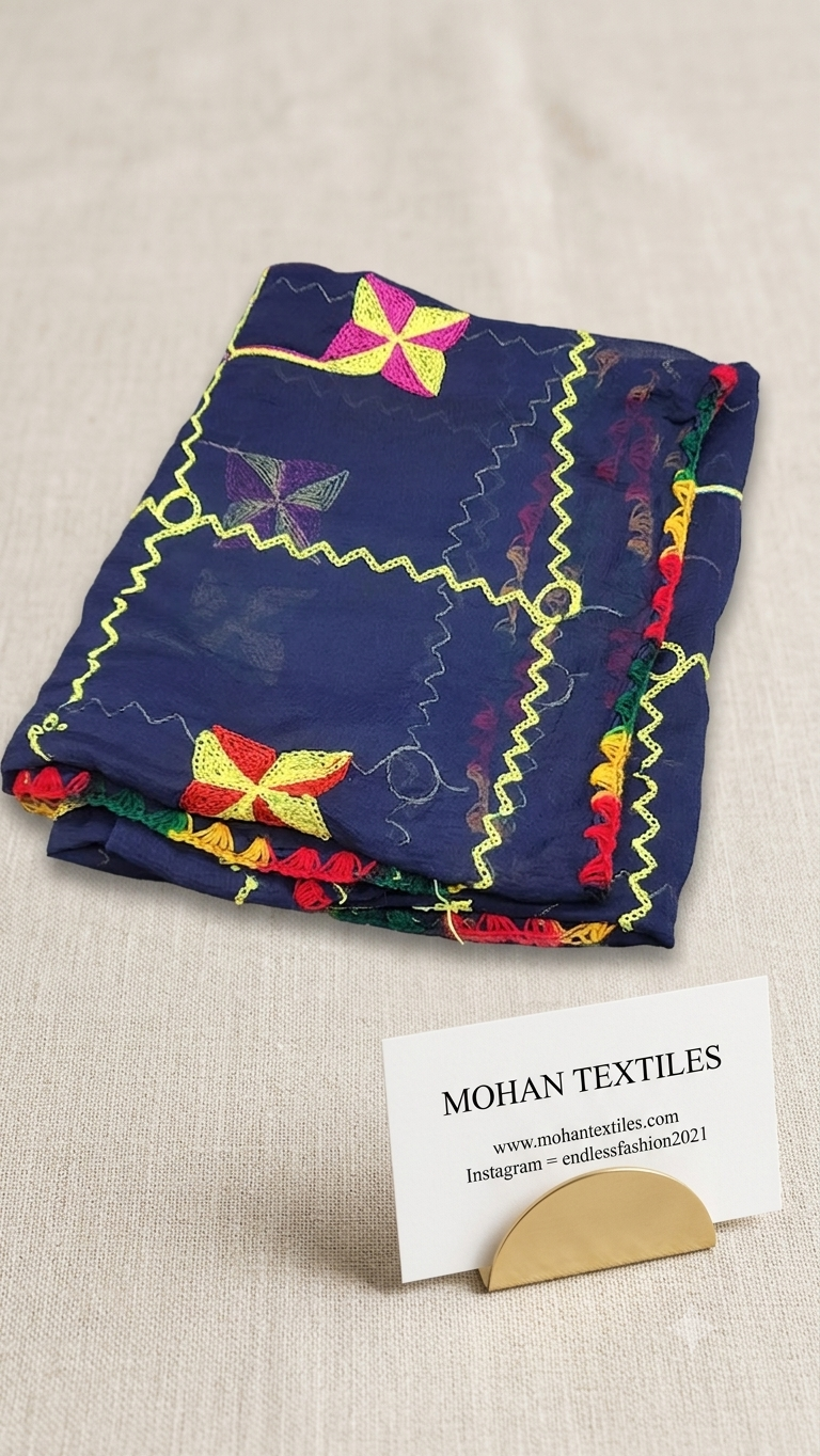 A folded navy blue fabric textile with colorful embroidered geometric and floral motifs, displayed on a light beige textured surface next to a business card for Mohan Textiles.