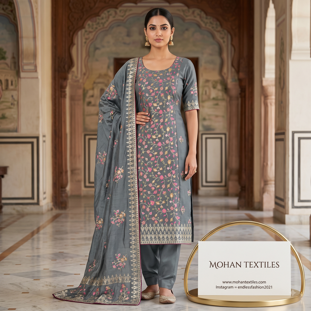 A woman models a grey floral Amritsari salwar kameez suit with a matching dupatta in an ornate indoor courtyard. The suit features pink and yellow floral embroidery, a patterned border, and matching grey trousers. Mohan Textiles.