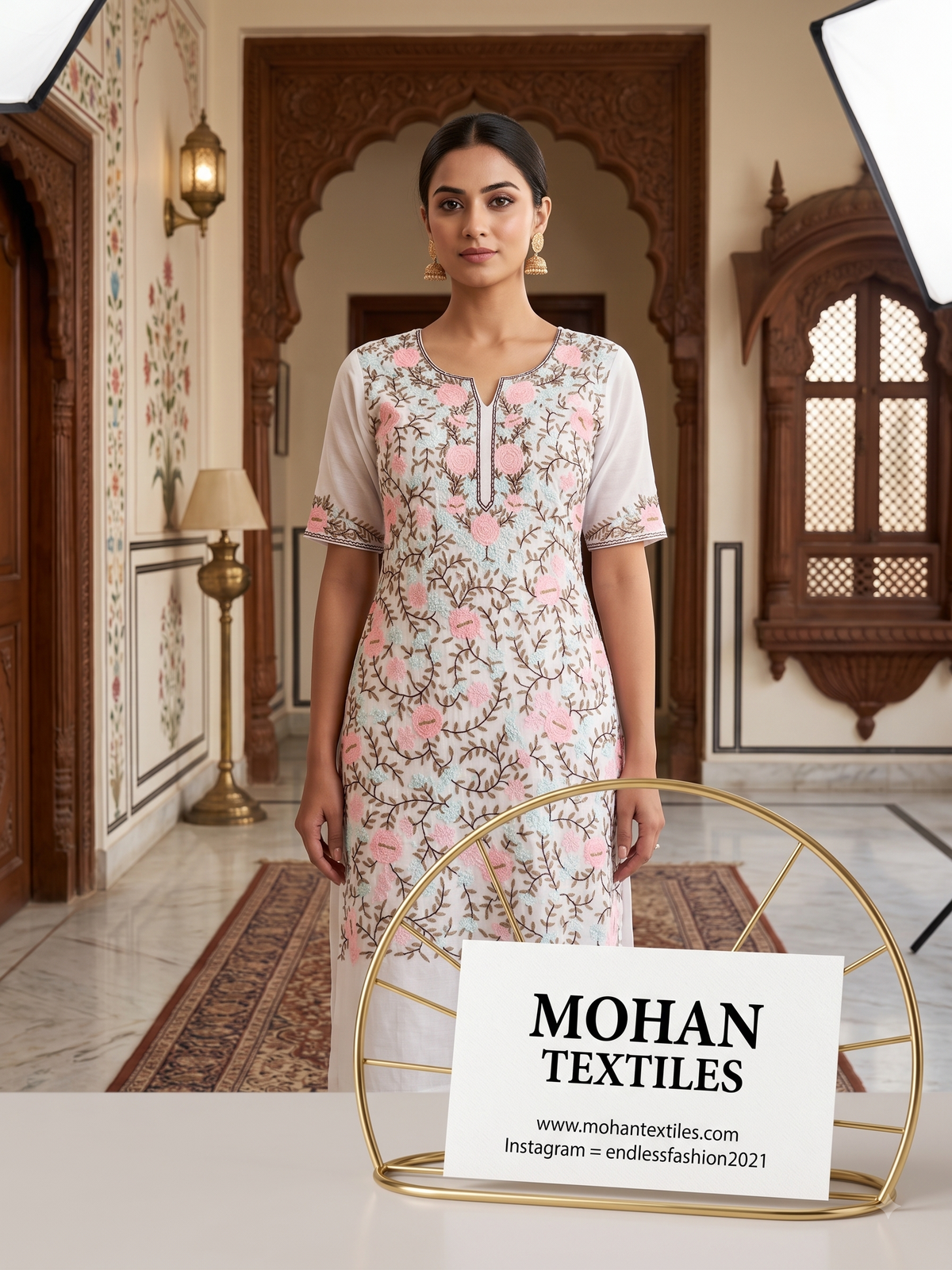 A woman stands indoors wearing a light pink and white floral Phulkari kurta with short sleeves and a keyhole neckline. A sign reading MOHAN TEXTILES is displayed in the foreground on a gold stand, with an ornate interior hallway behind her.