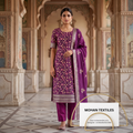 A woman stands in an ornate architectural corridor wearing a purple embroidered Amritsari salwar kameez set with matching pants and a dupatta. The outfit features floral embroidery and a decorative border by Mohan Textiles.