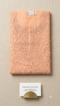 A folded peach-colored phulkari embroidered kurta or tunic with floral lace detailing, displayed on a textured beige fabric background. A small white brand card for 'MOHAN TEXTILES' is placed below the garment.