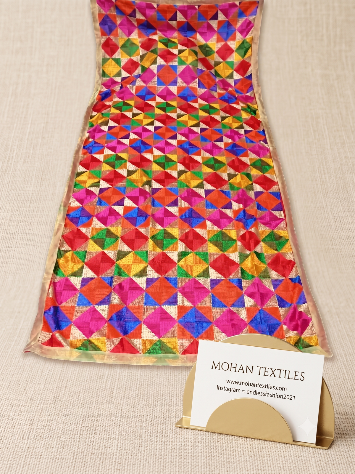 A colorful patterned fabric panel or scarf laid flat on a beige textured surface, with a small card holder displaying a business card for Mohan Textiles in front of it.