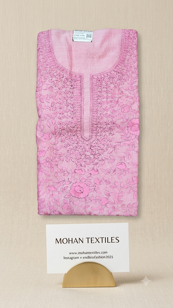 A folded light pink phulkari embroidered kurta or tunic with floral lace detailing, displayed on a beige surface with a card reading MOHAN TEXTILES and a small gold stand.