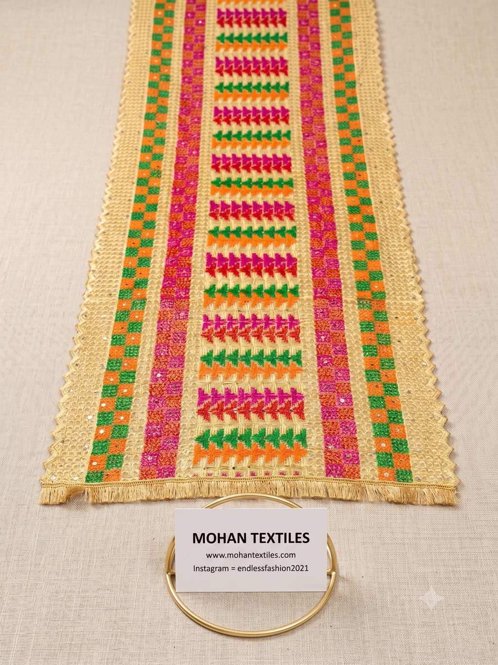 A close-up of a beige Phulkari Dupatta with colorful embroidered geometric patterns in pink, green, orange, and yellow, laid flat on a light fabric background with a small brand card and gold ring at the bottom.