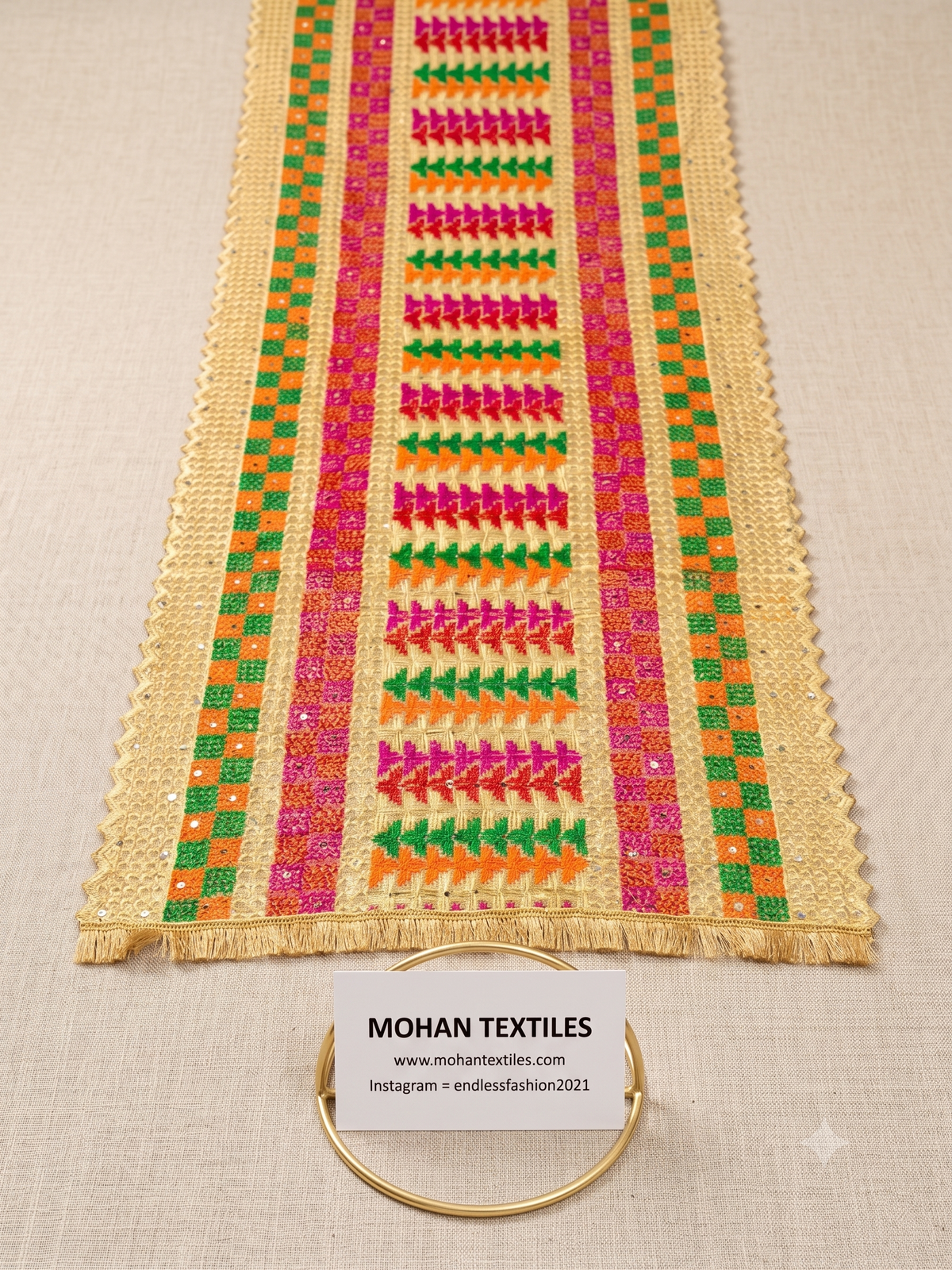 A close-up of a beige Phulkari Dupatta with colorful embroidered geometric patterns in pink, green, orange, and yellow, laid flat on a light fabric background with a small brand card and gold ring at the bottom.