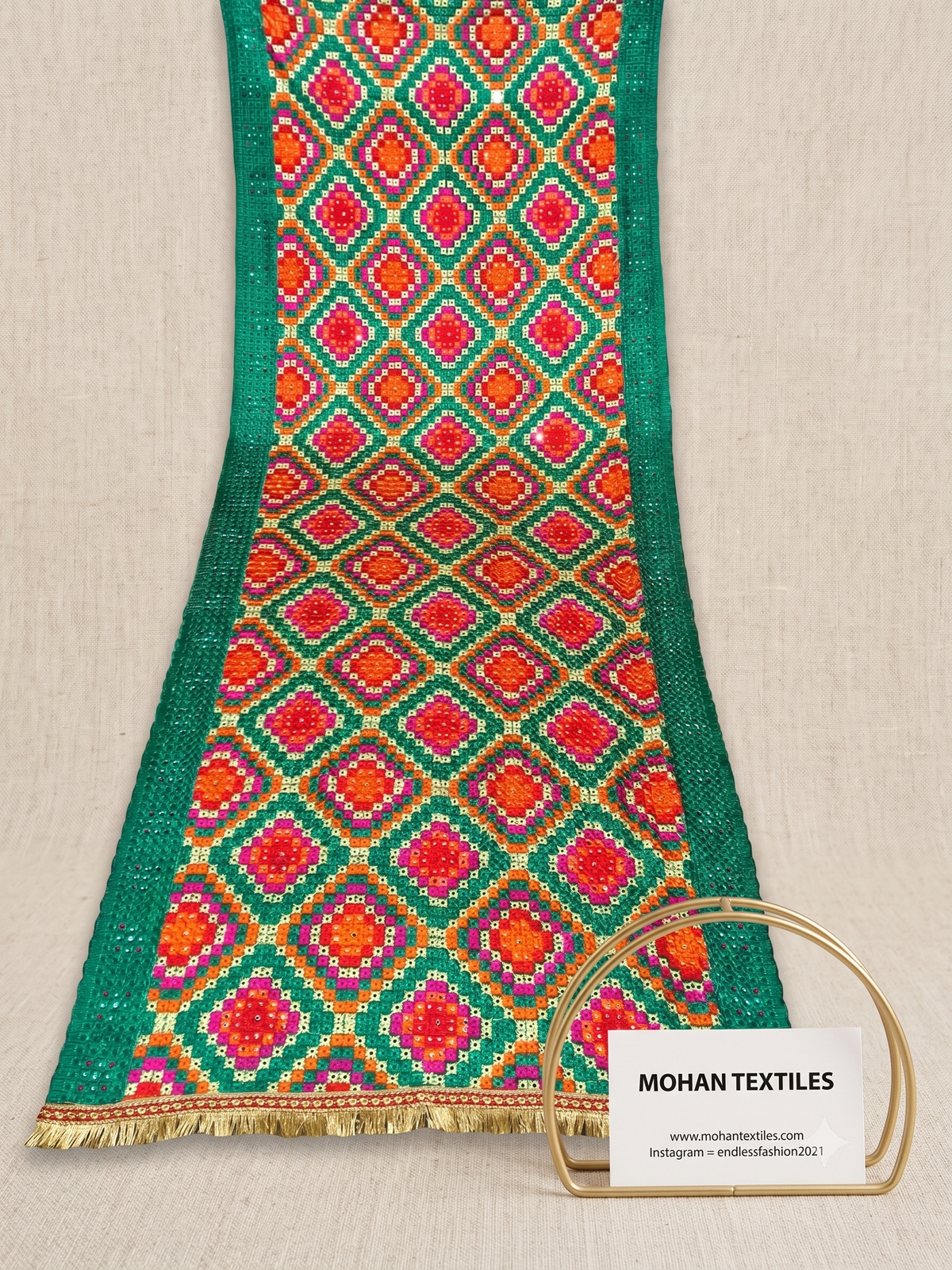 A close-up of a Phulkari Dupatta with a vibrant green border and a central diamond pattern in red, orange, yellow, and purple, finished with gold fringe. A small sign for Mohan Textiles is placed in front on a light beige fabric background.