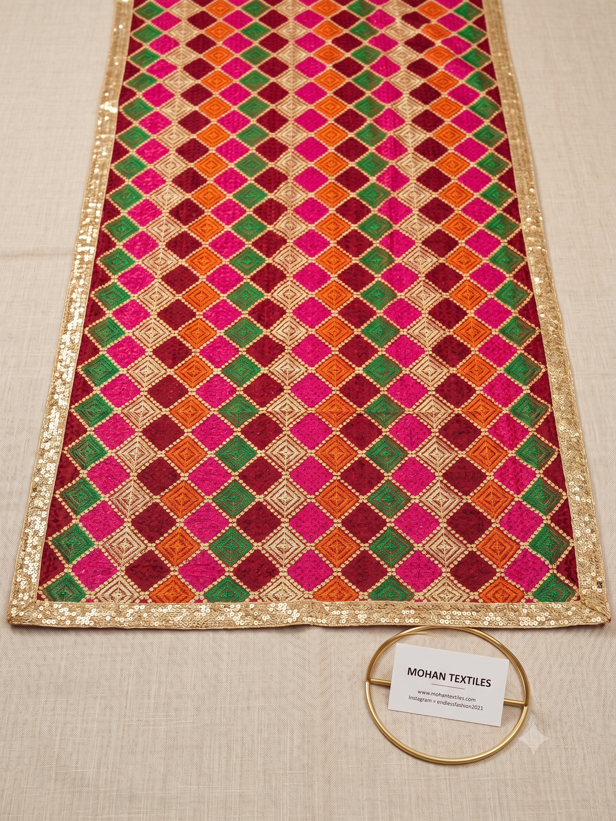A rectangular embroidered Phulkari Dupatta laid flat on a light beige fabric background. The design features a repeating diamond pattern in bright pink, green, orange, and deep red, bordered by metallic gold sequins.