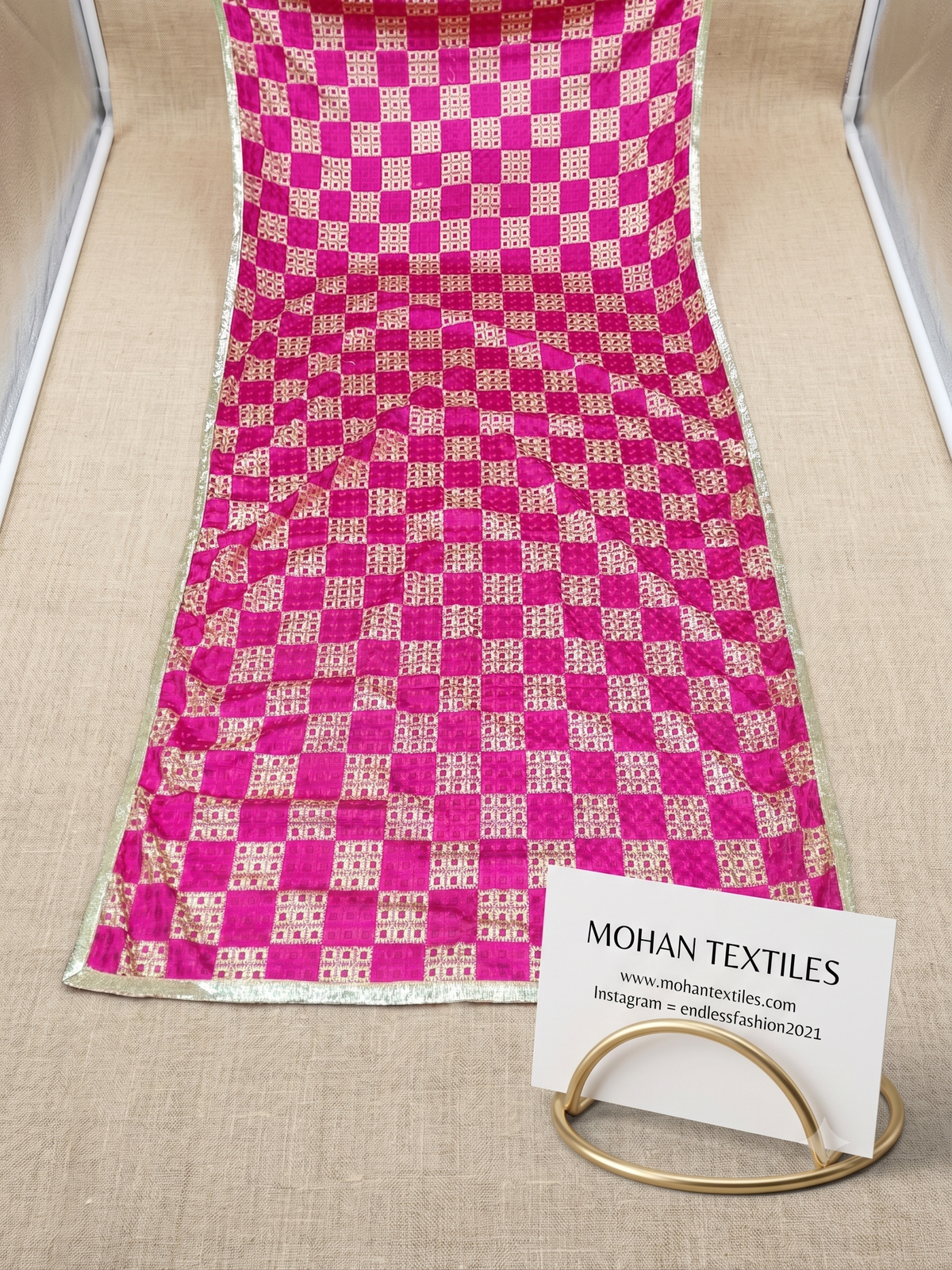A pink and gold Phulkari Dupatta Draped over a beige textured surface. A white card displaying the brand 'MOHAN TEXTILES' is positioned in the bottom right corner.