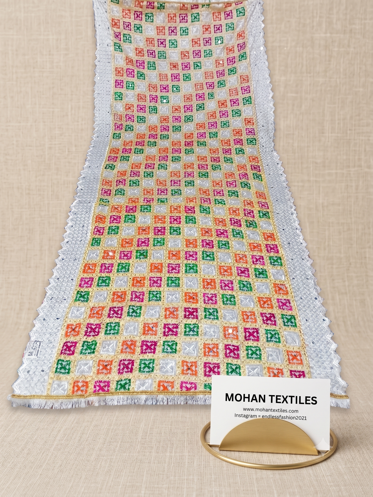 A long white Phulkari Dupatta with colorful embroidered checkered squares in pink, green, orange, and yellow, finished with white lace trim and fringe. A small card reading MOHAN TEXTILES is placed in front on a gold stand over a beige surface.
