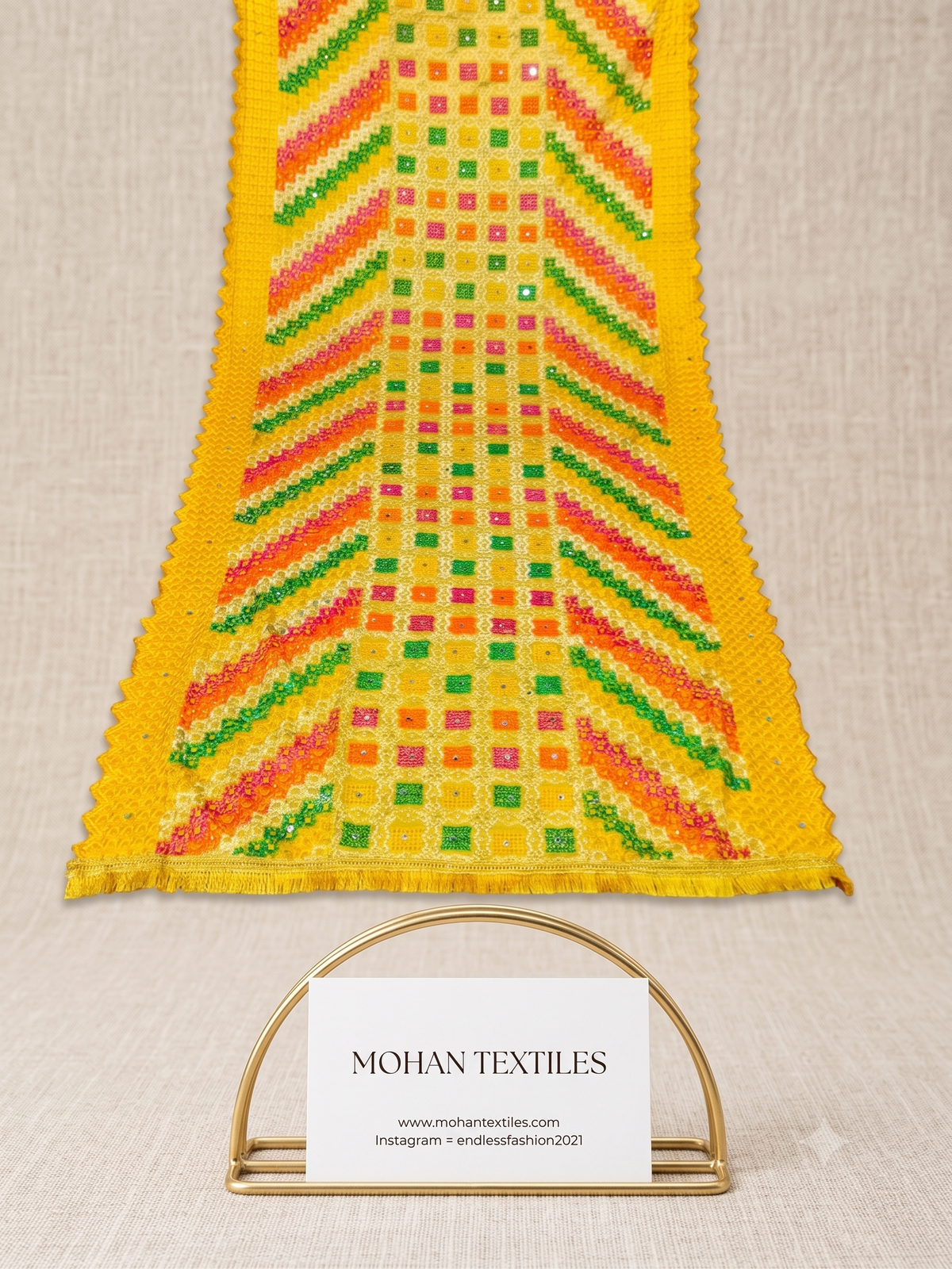 A close-up of a yellow Phulkari Dupatta with colorful geometric patterns in green, orange, pink, and red, displayed on a neutral textured background with a MOHAN TEXTILES brand card below.