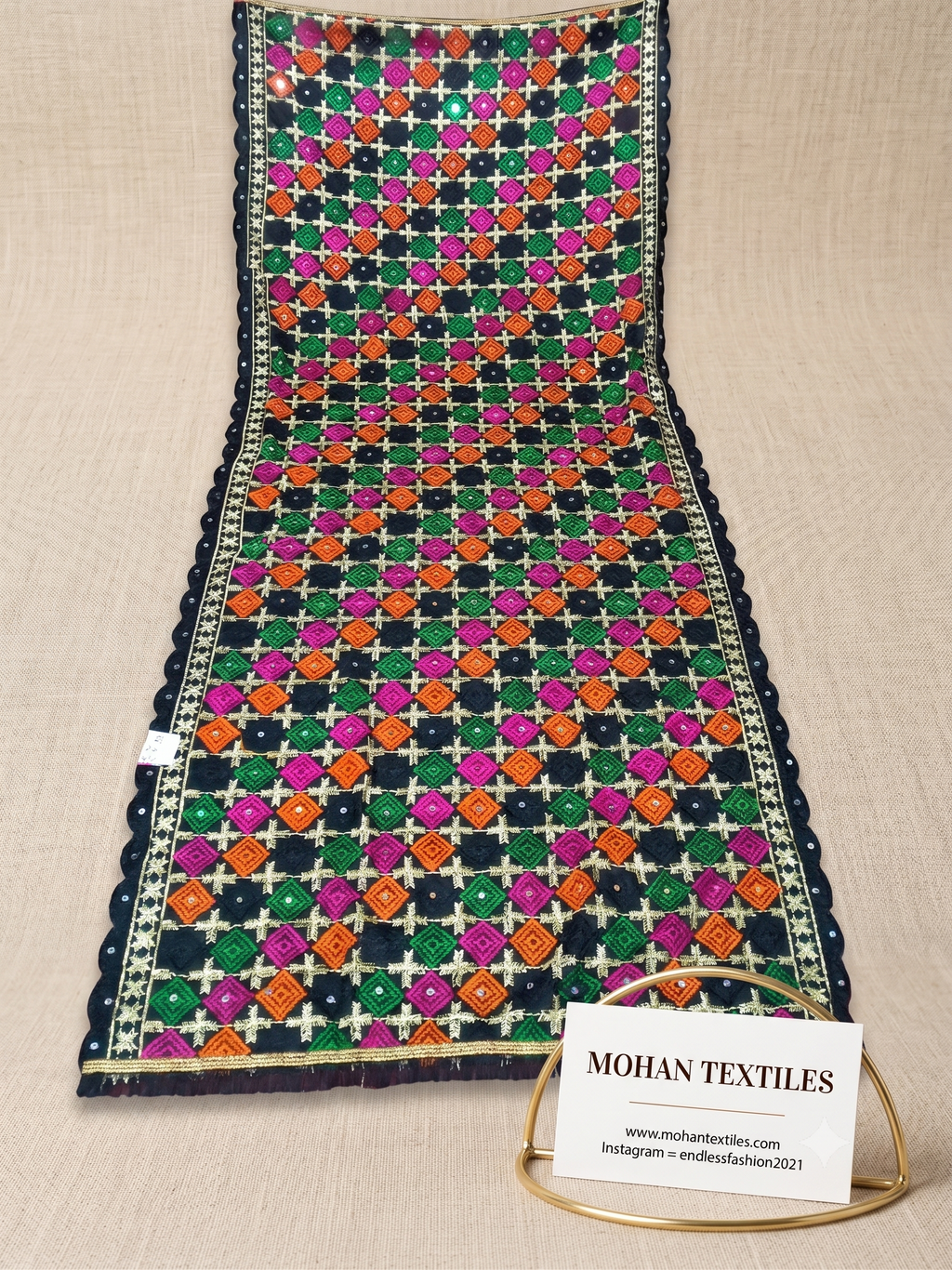 A folded and draped piece of Mohan Textiles Premium Phulkari Dupatta  featuring a vibrant multicolor ikat pattern with pink, green, orange, and black motifs on a cream base, displayed on a neutral beige textured background with a brand card.
