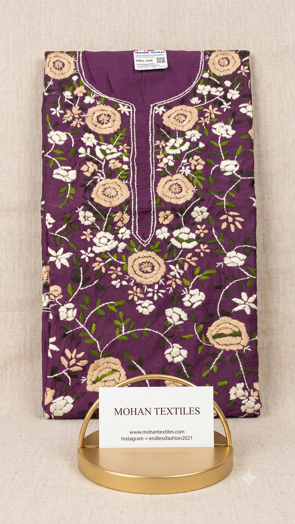 A folded purple phulkari embroidered kurta or tunic with floral and vine motifs, displayed on a gold stand with a white card labeled MOHAN TEXTILES. The fabric is laid flat on a light beige textured background.