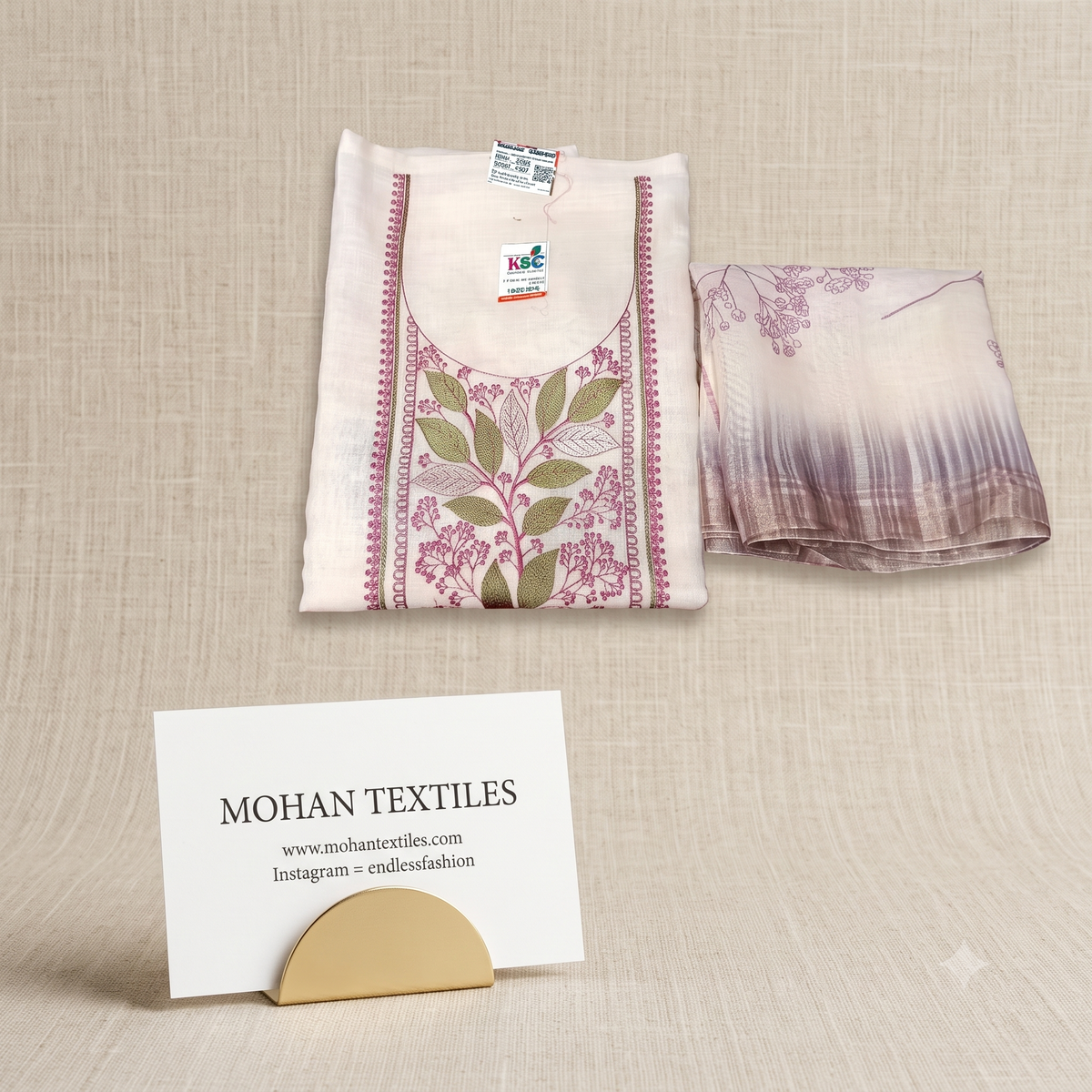Overhead view of a folded off-white linen unstitched suit kurta with vibrant magenta and green leaf thread embroidery, next to a matching floral printed dupatta. Quality linen fabric from Mohan Textiles.
