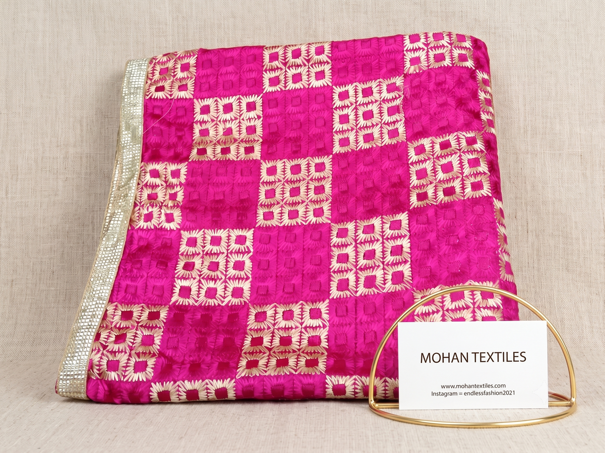 A folded pink and white patterned phulkari dupatta fabric, displayed on a neutral beige fabric background. A small white card with the brand name 'MOHAN TEXTILES' is held in a gold wire stand in the foreground.