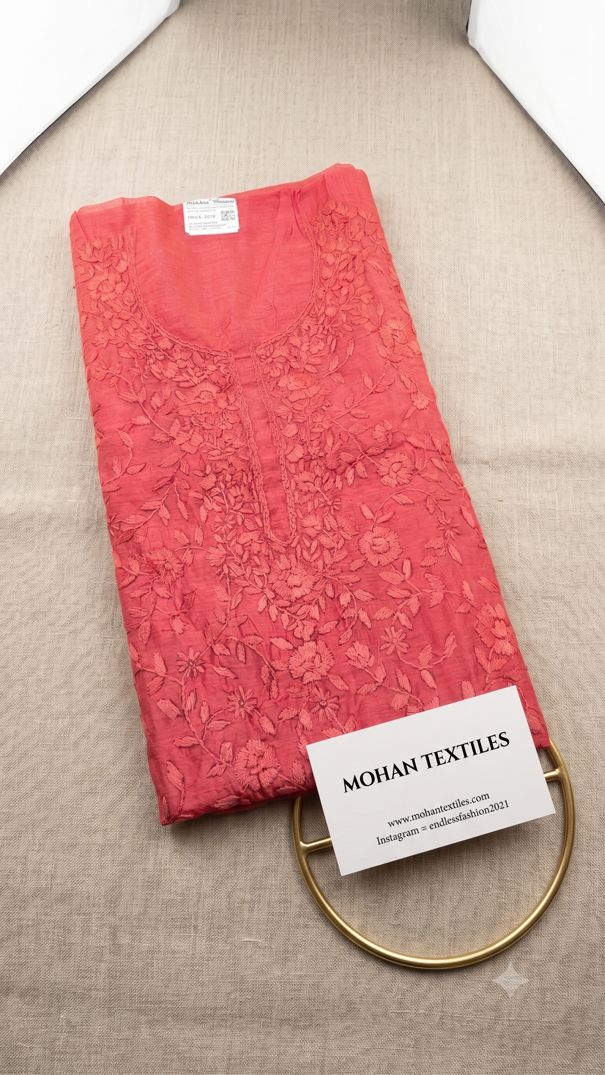 A folded coral pink phulkari embroidered kurta fabric, displayed on a beige textured fabric surface with a white brand card reading MOHAN TEXTILES and a gold ring stand.