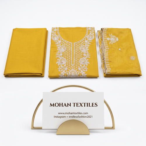 Three folded pieces of mustard yellow fabric displayed on a white background, with the center piece featuring white floral embroidery and the right piece showing embroidered borders. A gold metal stand holds a white card reading MOHAN TEXTILES.