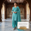 A woman stands in a grand, ornate indoor corridor wearing a turquoise embroidered salwar kameez suit with matching pants and a draped dupatta. A white sign in the lower right reads 'MOHAN TEXTILES' with website and Instagram details.