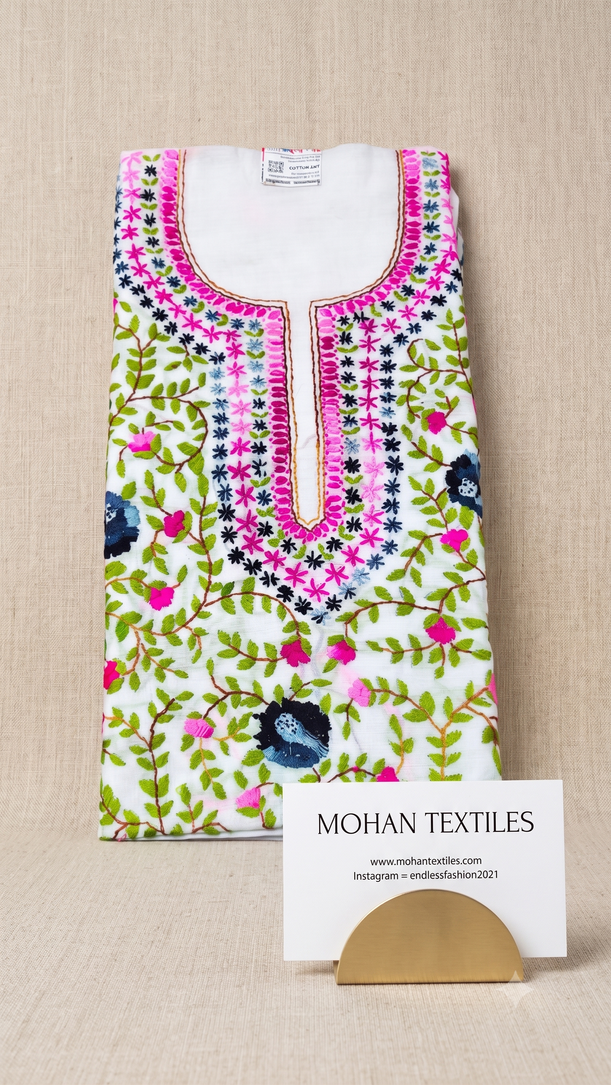 A folded white kurta with vibrant pink, green, and blue floral embroidery, displayed on a beige textured background with a Mohan Textiles business card.