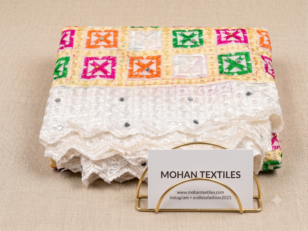 A folded phulkari dupatta of Mohan Textiles featuring a yellow and white geometric embroidered pattern on the top half and a white lace border on the bottom half, displayed on a beige fabric background with a gold stand and brand card.